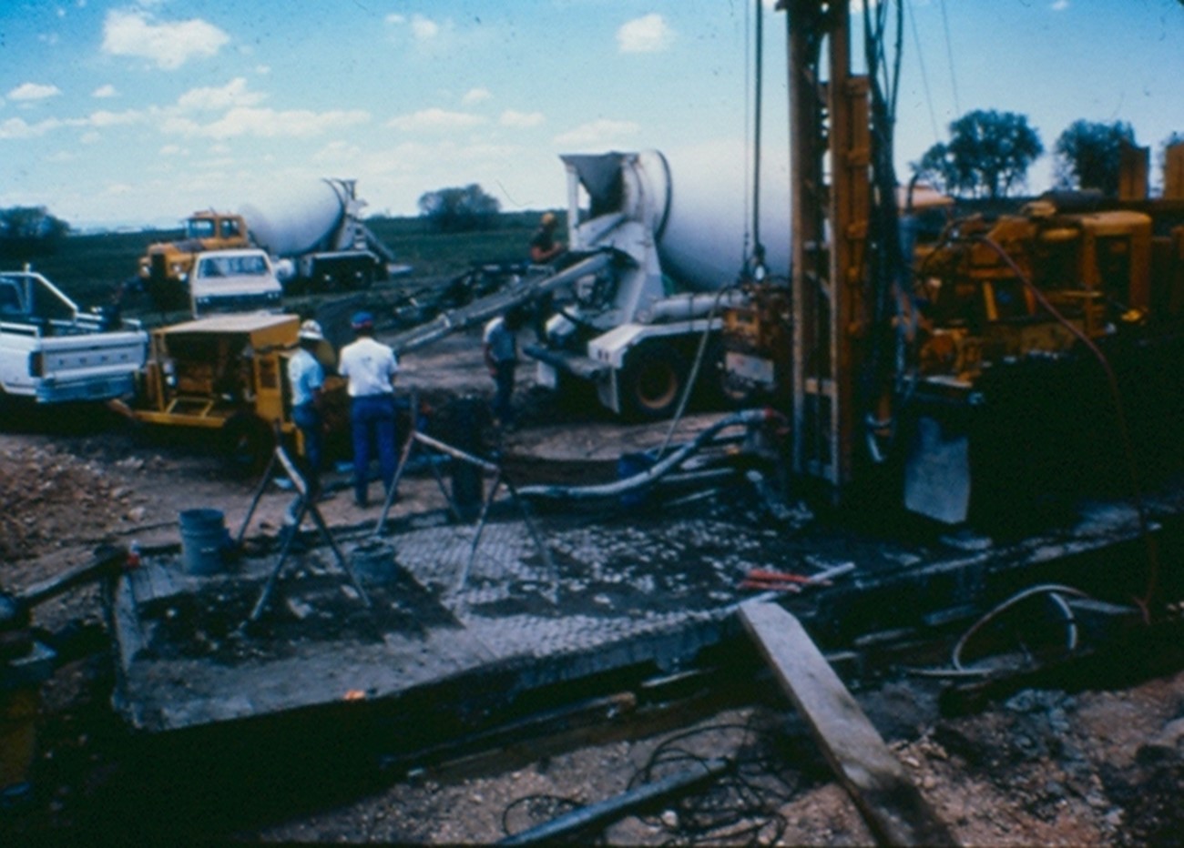 This image shows the installation of a grout wall to prevent the spread of an underground mine fire.  There is a drill rig which bores a hole to the burning coal seam.  Then a grout pump is set to pump grout into the mine voids sealing them in the hopes that the fire will be blocked.  It can take many, many tons of grout to create the barrier wall.