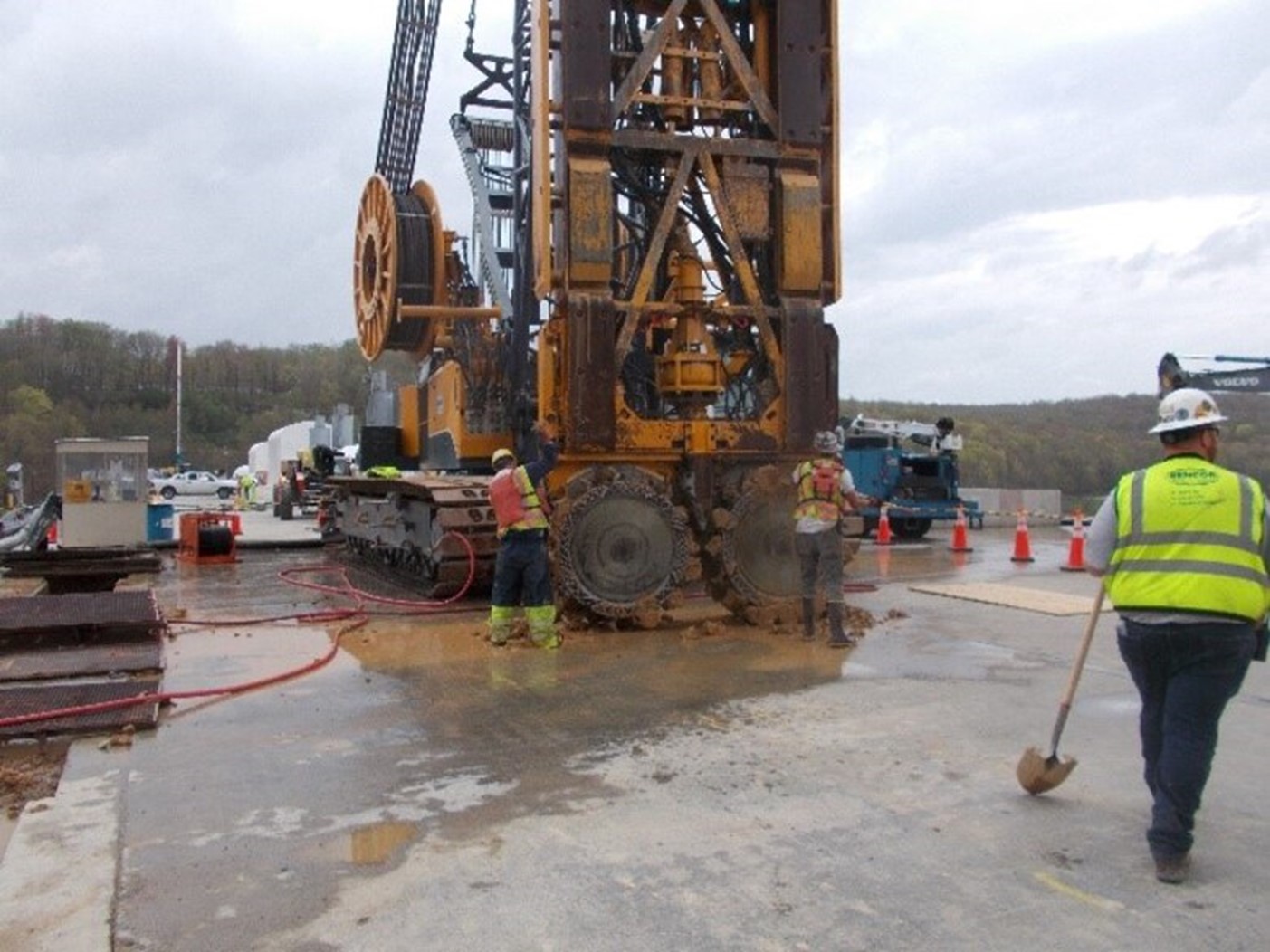 A photo by Greg Hensley, shows a piece of equipment which can dig a trench 3 feet by 10 feet to a predetermined depth.  There are two large cutting wheels attached to a crane-like tracked equipment.  The image shows workers cleaning the cutting heads after completing a vertical trench cut.