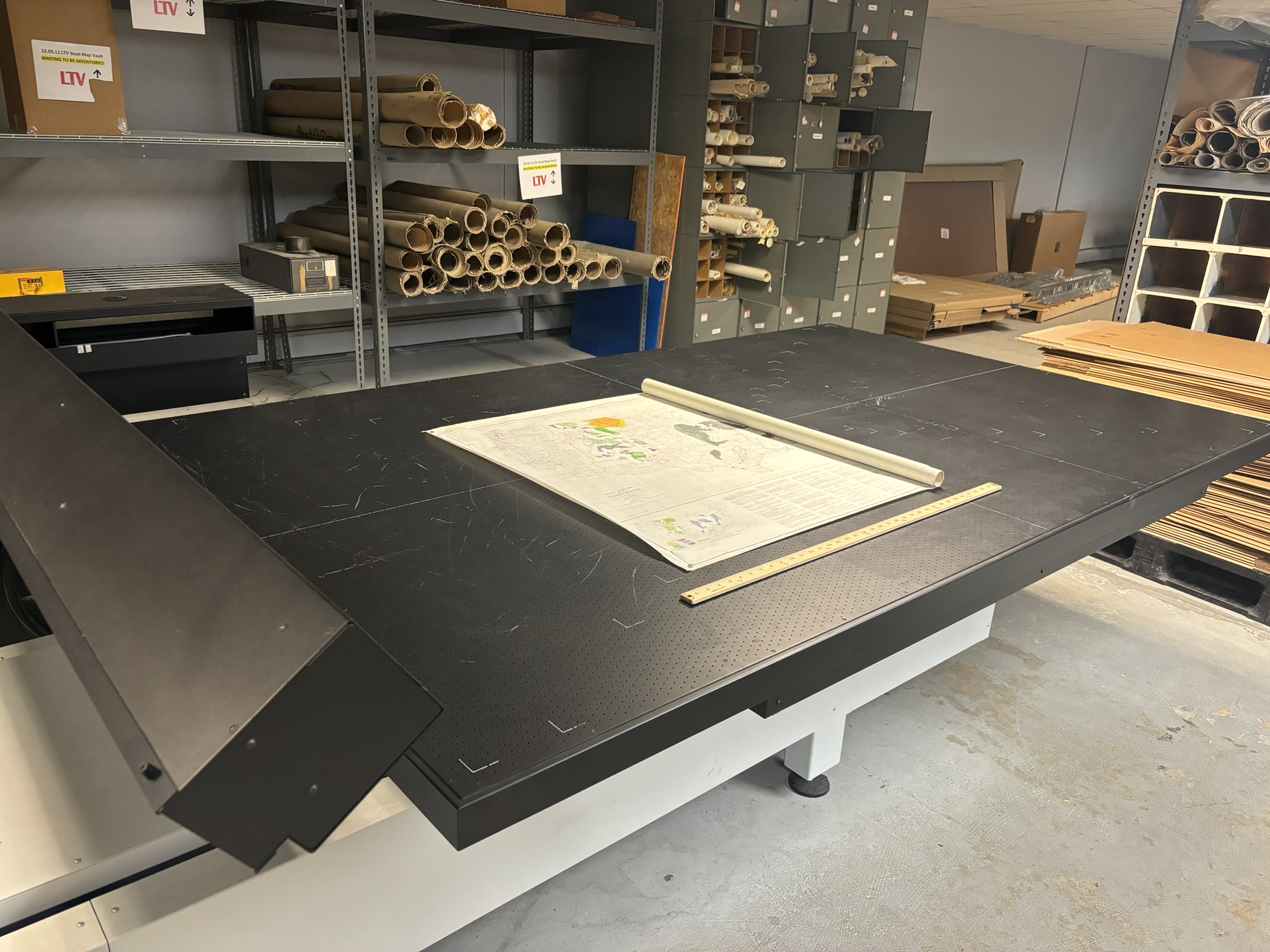 Photo of large flatbed scanner with a map halfway unrolled on top.  Rolled maps visible in background.