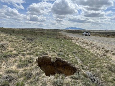 A hole in the ground sits to the left of a dirt road under light blue sky and puffy white clouds. A truck drives away and toward mountains in the distance.