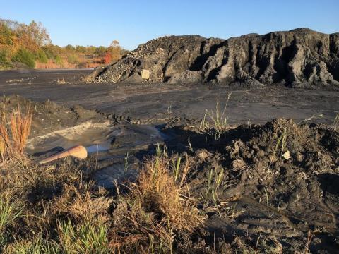 A rusted pipe is exposed against wet gray and black dirt of a tall coal waste pile in the background. Sunny blue sky and fall trees line the far background.