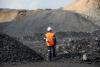 A male in orange safety vest and white hardhat stands with his back to the camera, in the midst of and overlooking tall, black coal waste piles